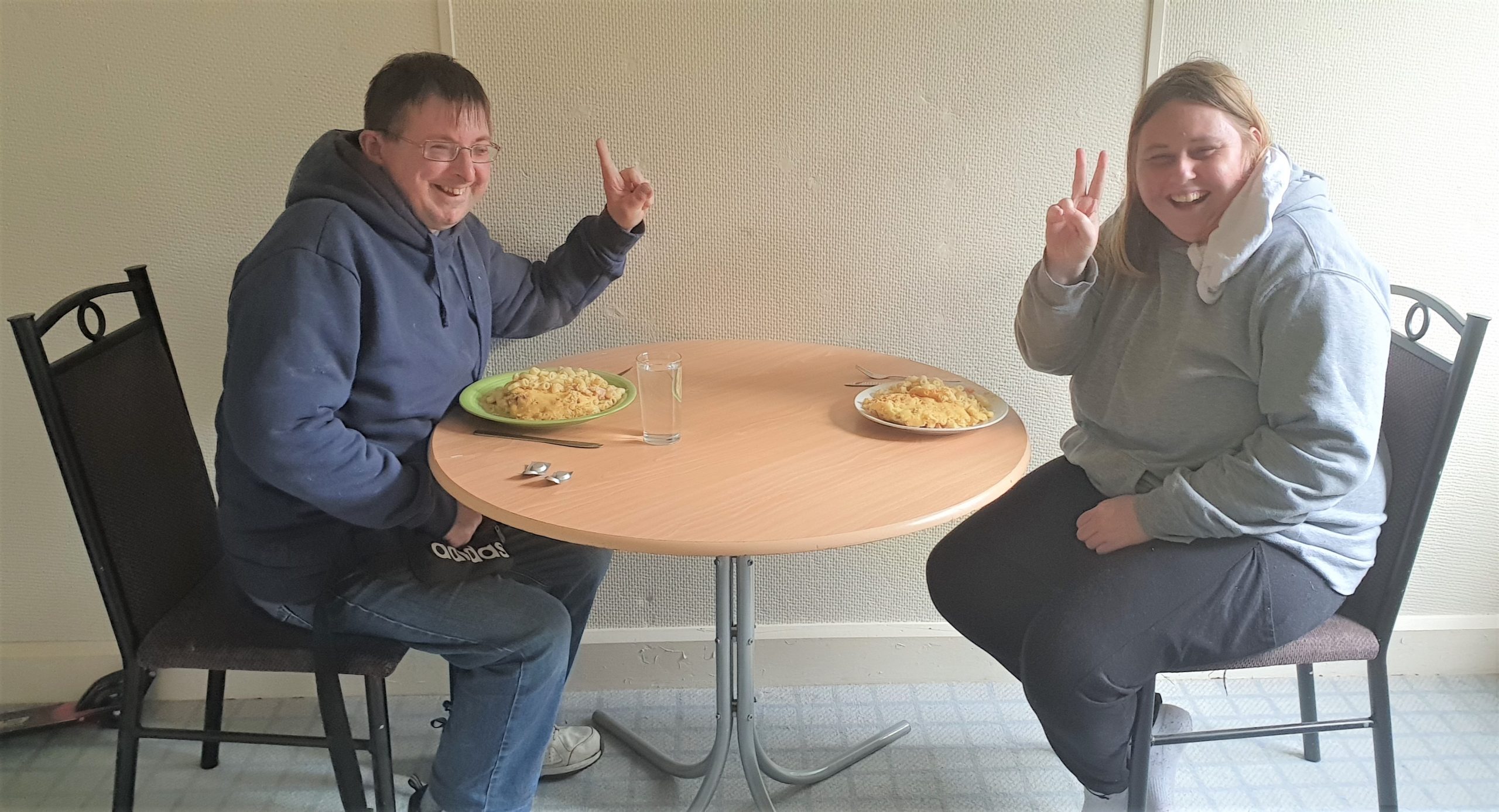 Jordan and Courtney Jordan (left) and Courtney (right) sitting down at a table holding the peace sign up. They are both smiling with a pasta dish on the table.